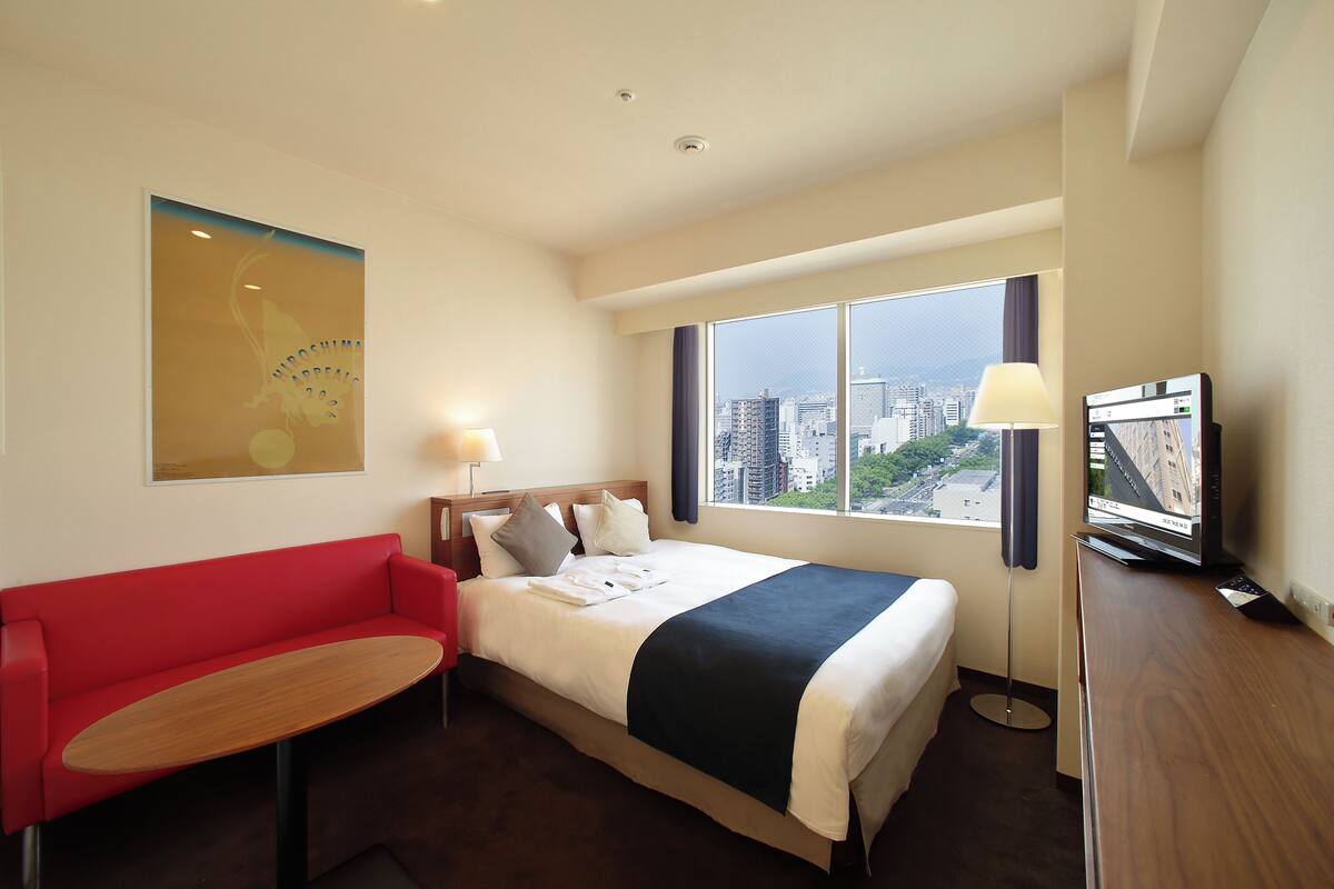 Habitación de hotel con cama, sofá rojo, mesita, televisor y ventana grande con vista a la ciudad.