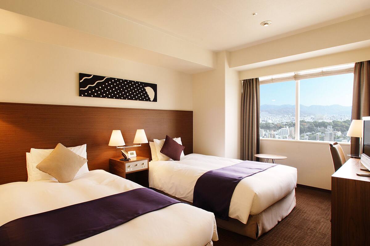 Habitación de hotel con dos camas, una mesita de noche con lámparas, un escritorio con una silla y un amplio ventanal con vistas a la ciudad.