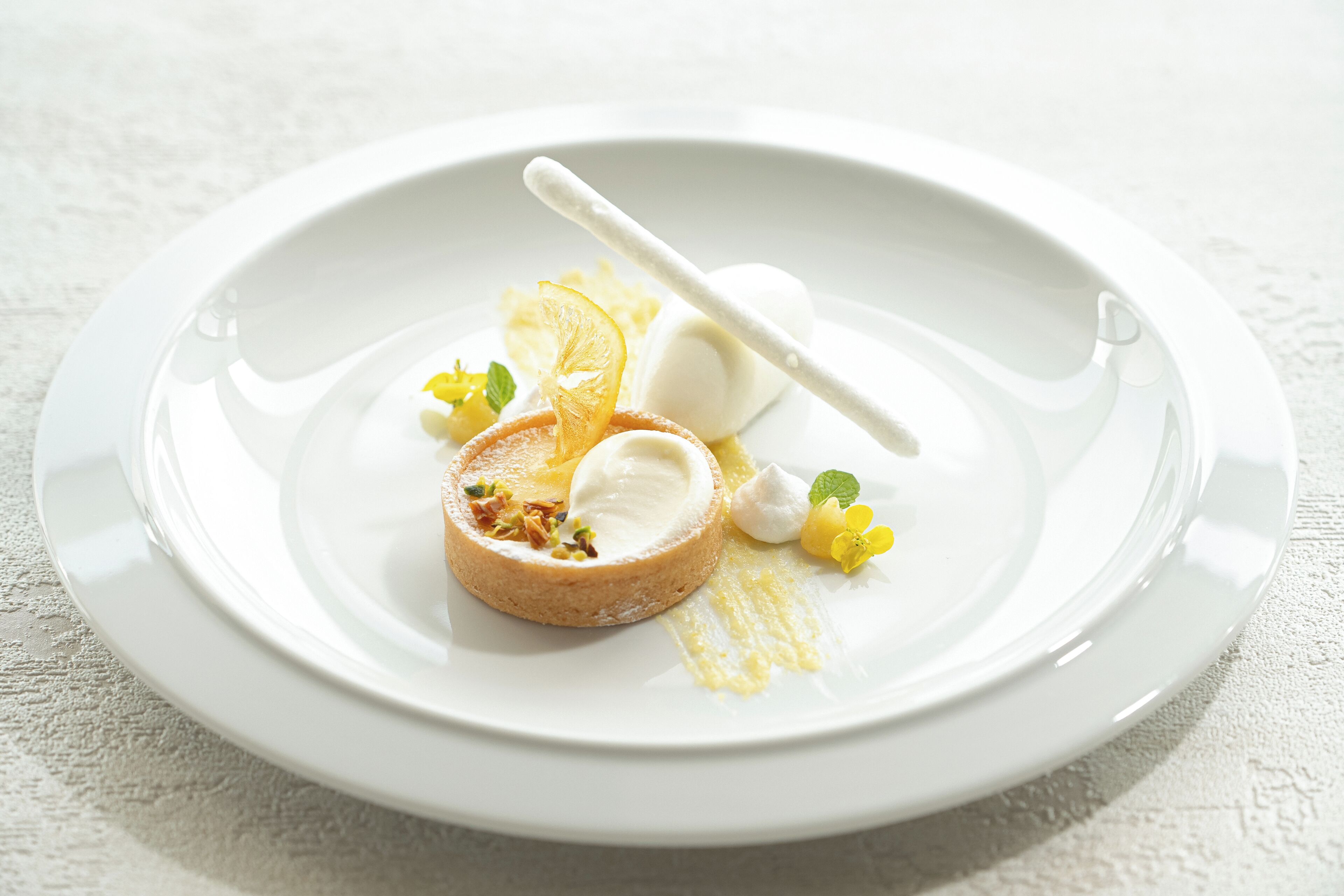 Une assiette blanche contenant un dessert composé d’une pâtisserie ronde, d’une boule de glace et d’un bâtonnet blanc, le tout décoré de fleurs jaunes et de tranches d’orange.