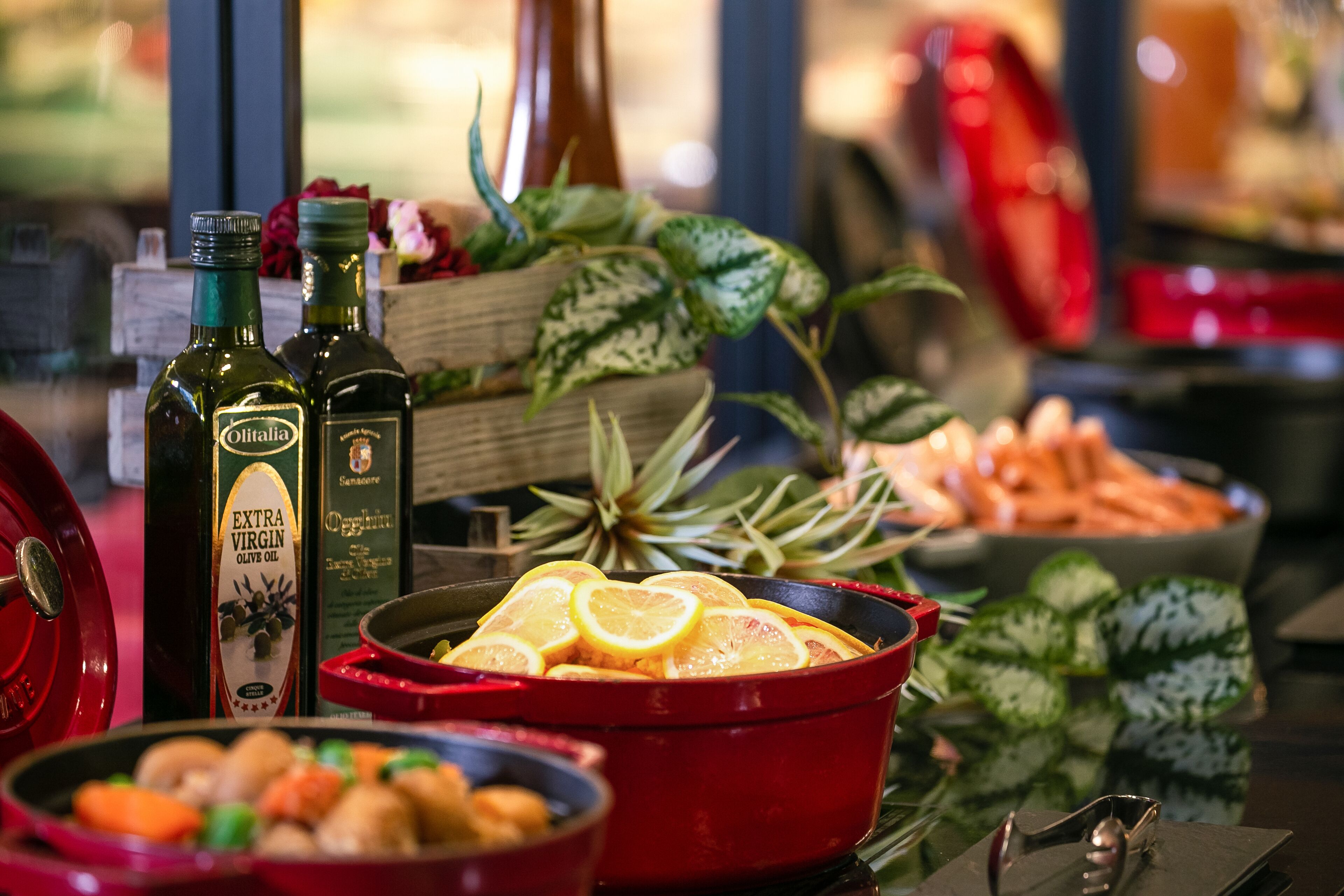 A table with a red pot containing sliced lemons, two bottles of olive oil, and a bowl of vegetables.