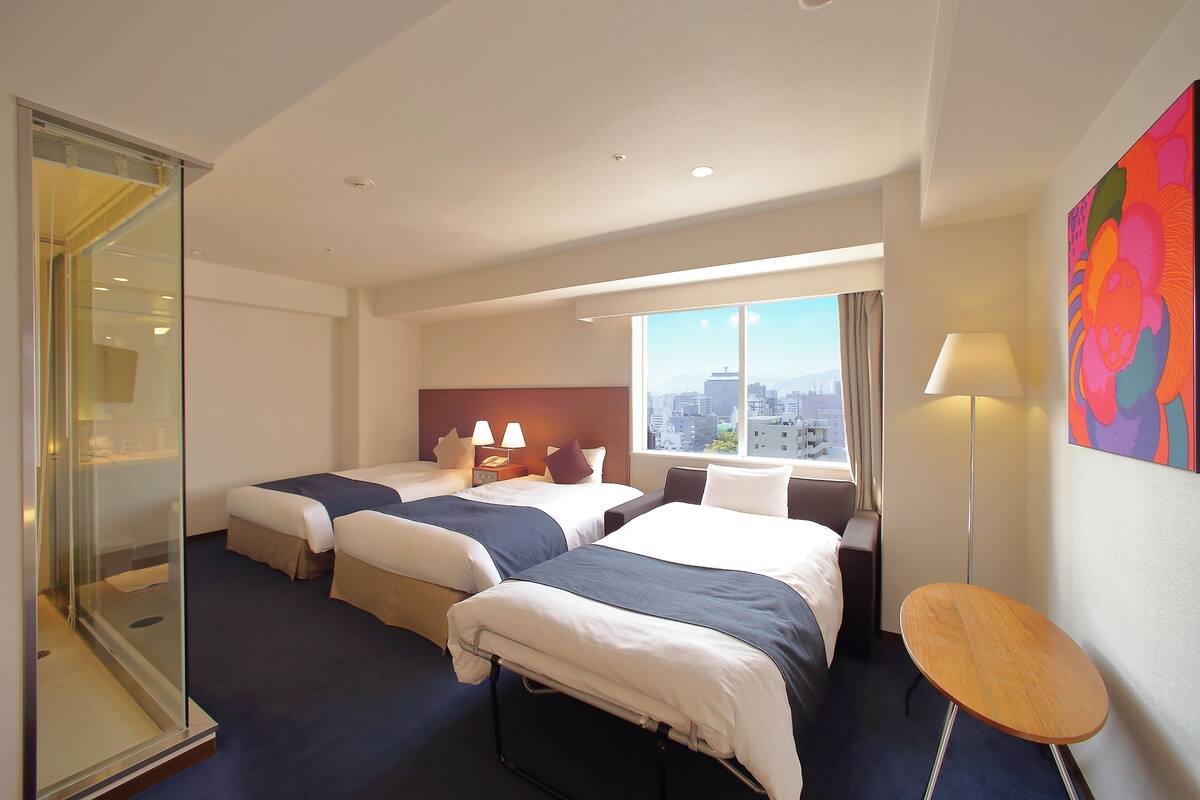 Habitación de hotel con dos camas, una mesita pequeña y un ventanal amplio con vistas a la ciudad.