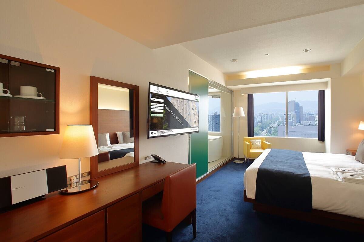 Habitación de hotel con cama, escritorio, silla, espejo, televisor y ventana con vista a la ciudad.