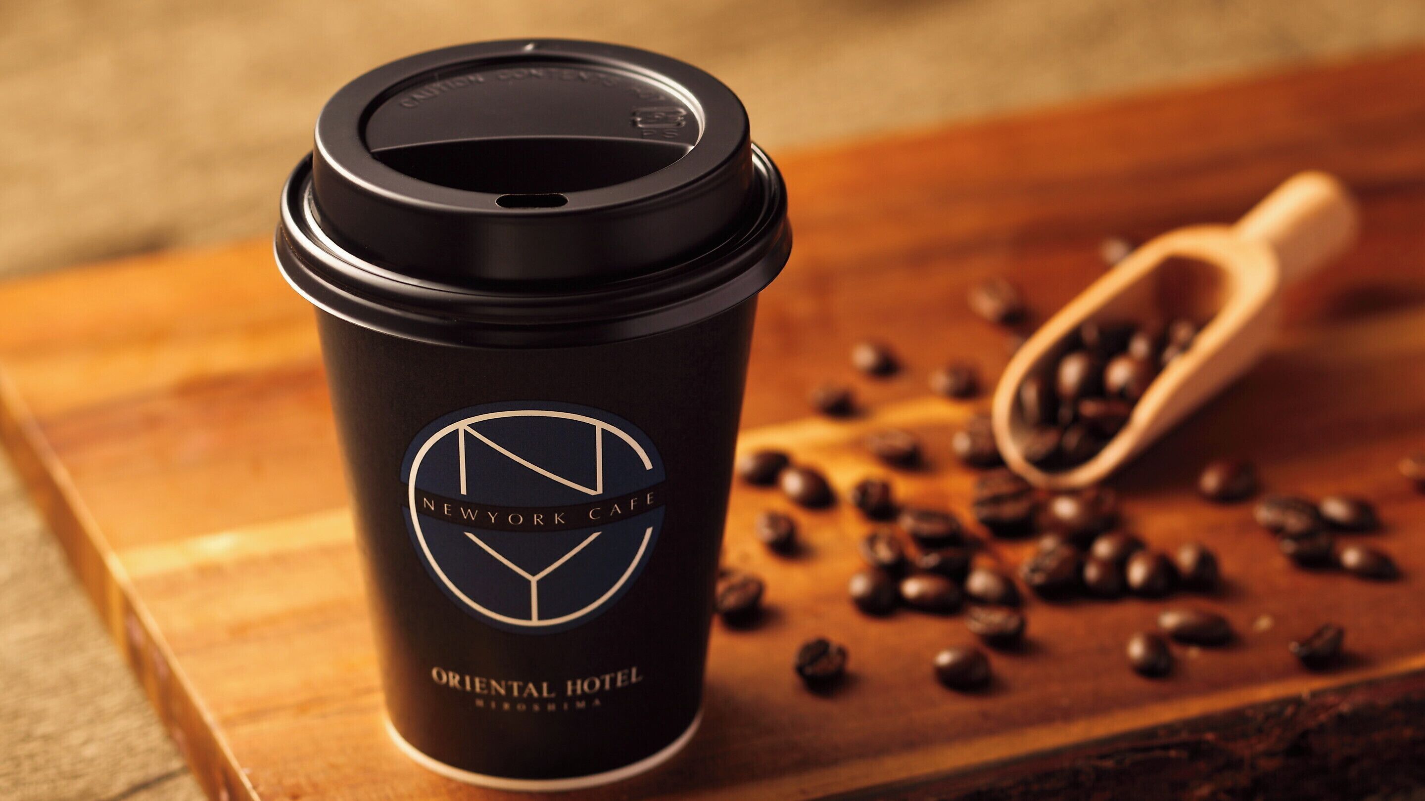 A black coffee cup with a lid and a logo, placed on a wooden surface with scattered coffee beans and a wooden scoop.
