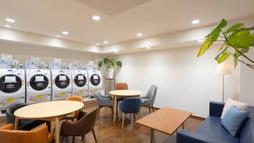 A laundry room with multiple washing machines, a table, chairs, a sofa, and a potted plant.