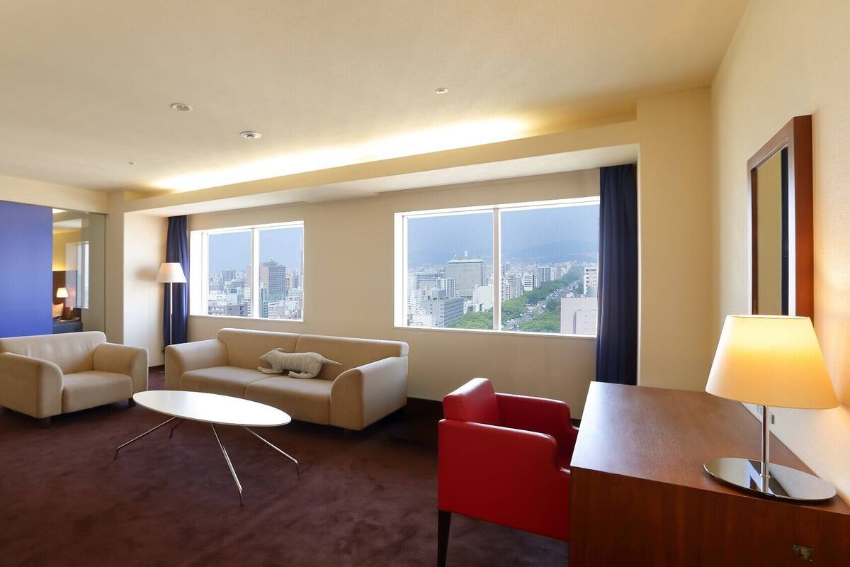 Habitación de hotel con sofá, dos sillones, una mesita, escritorio con lámpara y ventanas amplias con vistas a la ciudad.