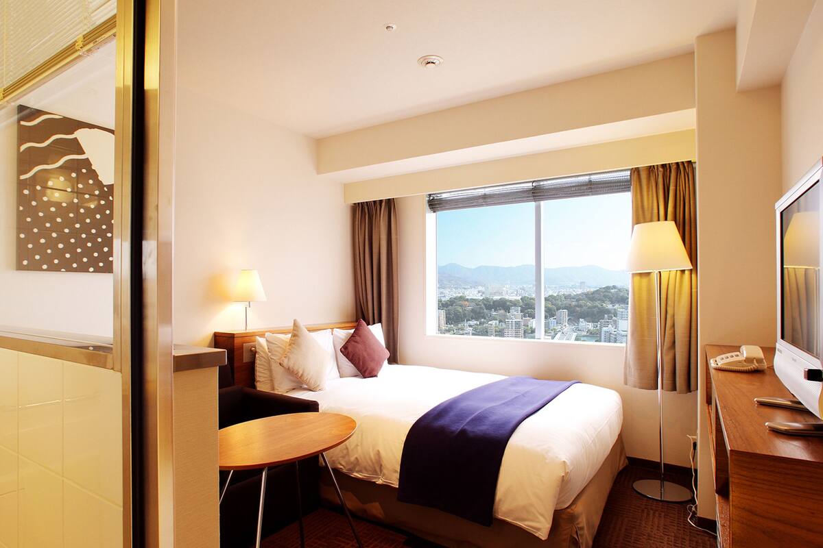 Habitación de hotel con cama, mesita de noche, lámpara, televisor y vista a un paisaje urbano por la ventana.