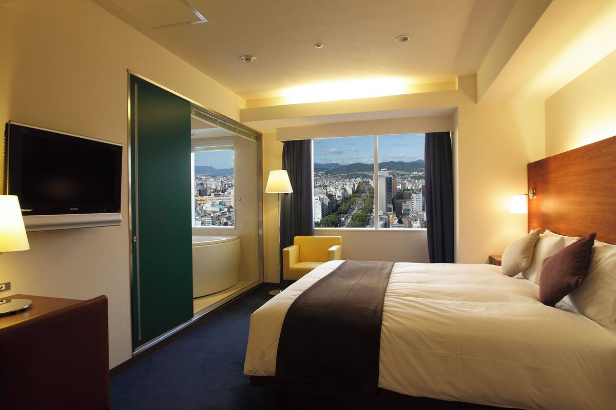Una habitación de hotel con una cama, televisión, una silla y un ventanal con vistas a la ciudad.