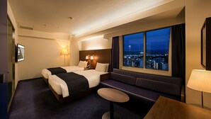 Habitación de hotel con dos camas, un sofá, una pequeña mesa redonda y un ventanal con vistas a la ciudad.