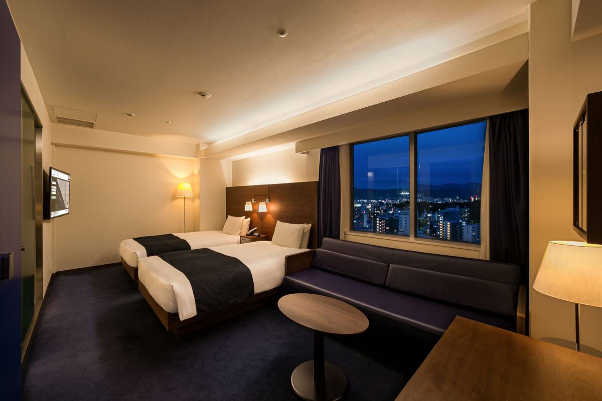 Habitación de hotel con dos camas, un sofá, una pequeña mesa redonda y un ventanal con vistas a la ciudad.