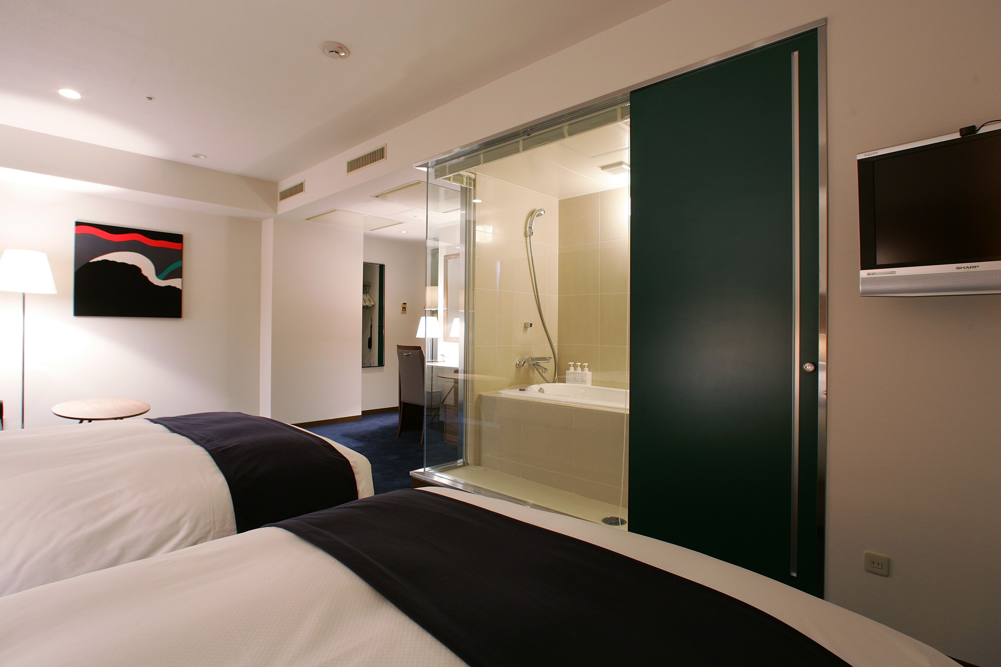 Habitación de hotel con dos camas, zona de ducha con paredes de vidrio, televisor montado y un cuadro en la pared.