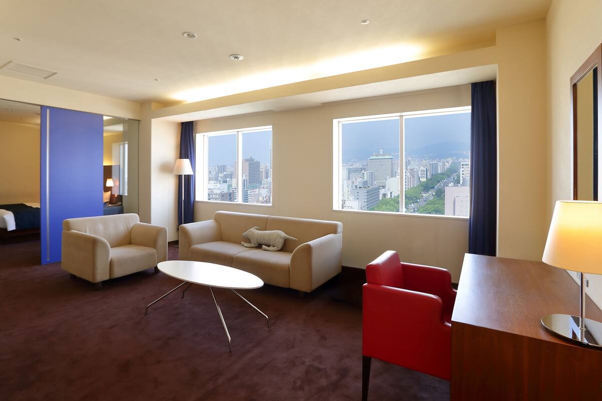Habitación de hotel con sofá, sillón y una mesita. Amplias ventanas ofrecen vistas a la ciudad.