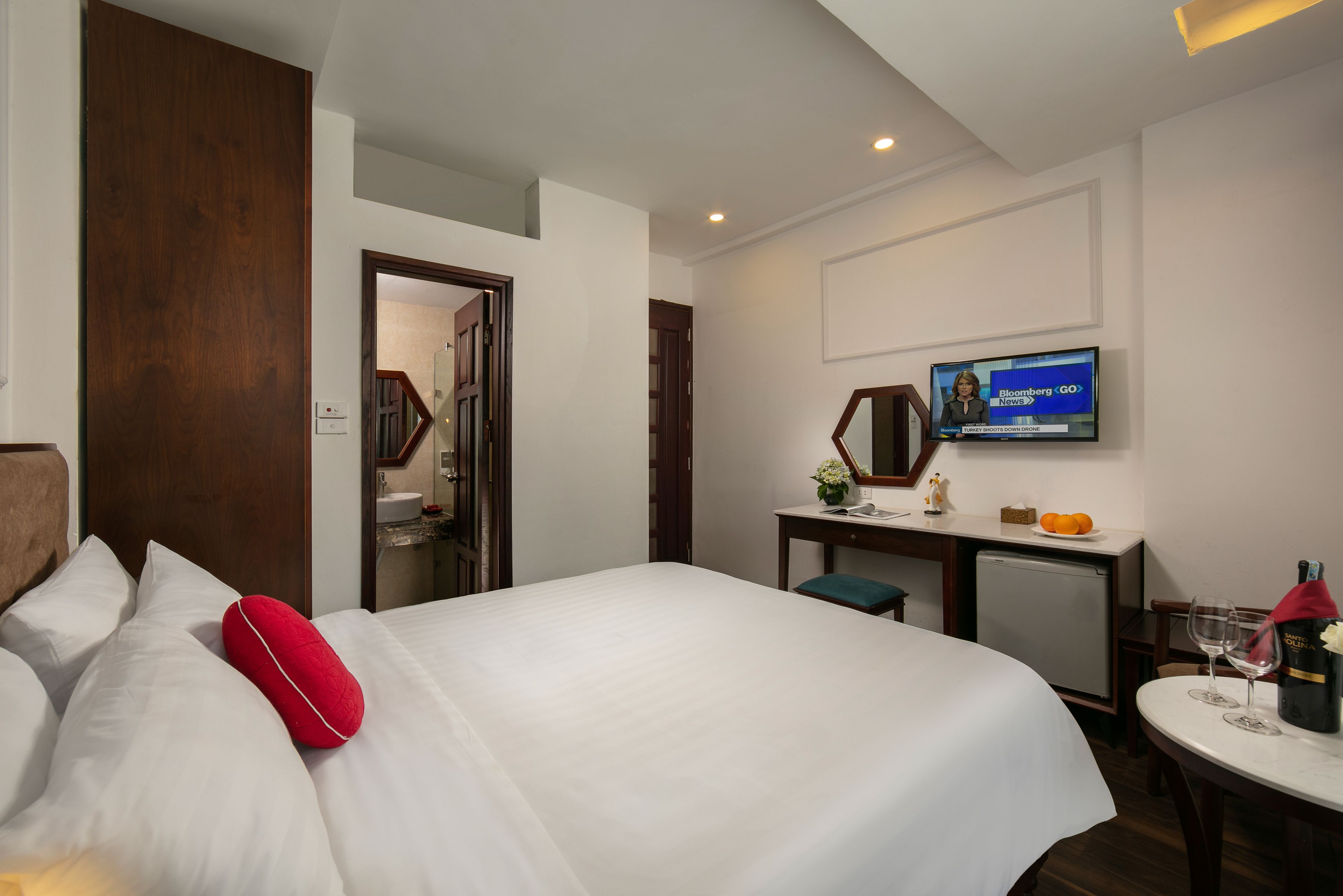 A hotel room with a large bed, a desk with a television, a small refrigerator, and a dining table with wine glasses.