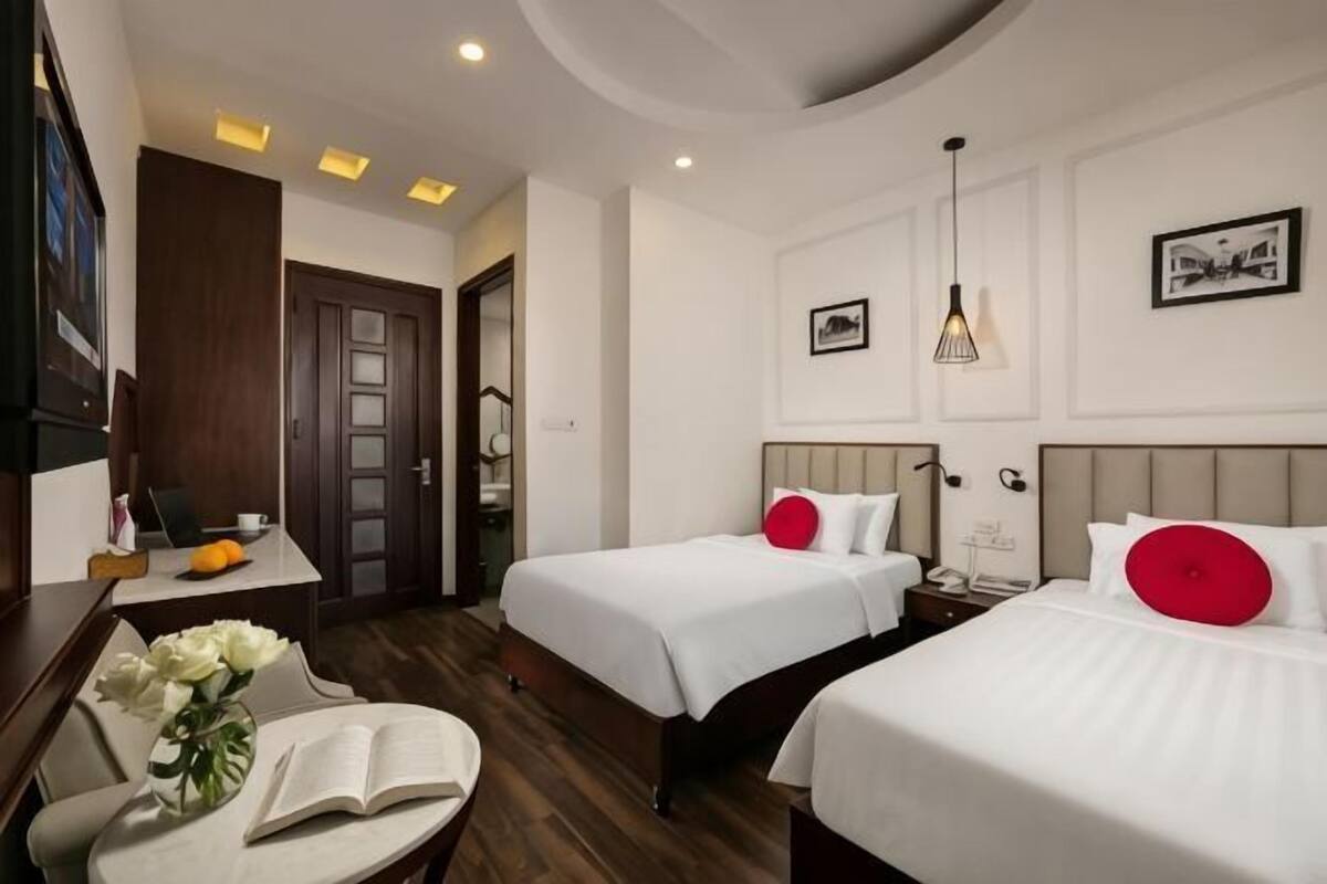 A hotel room with two beds, a desk, a chair, a vase with flowers, and a bookshelf.