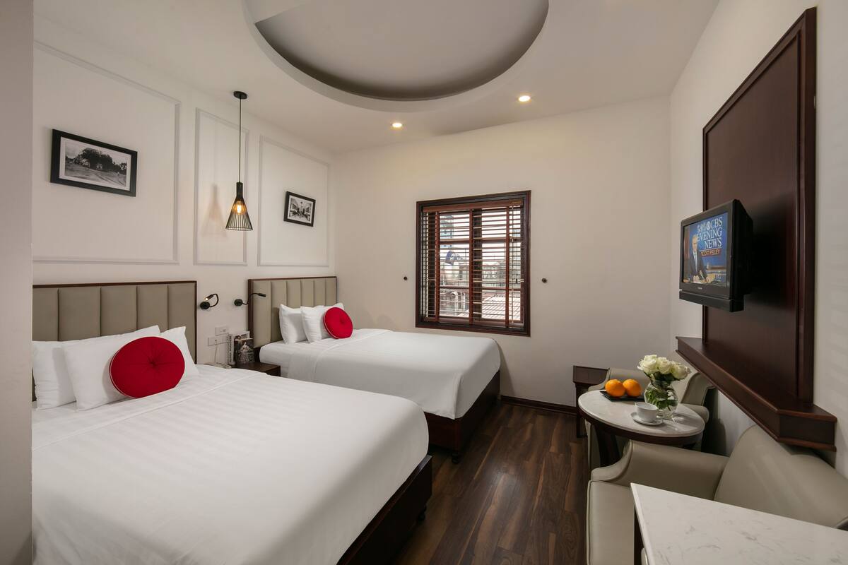 A hotel room with two beds, a small table with a vase of flowers, a television, and a window with shutters.
