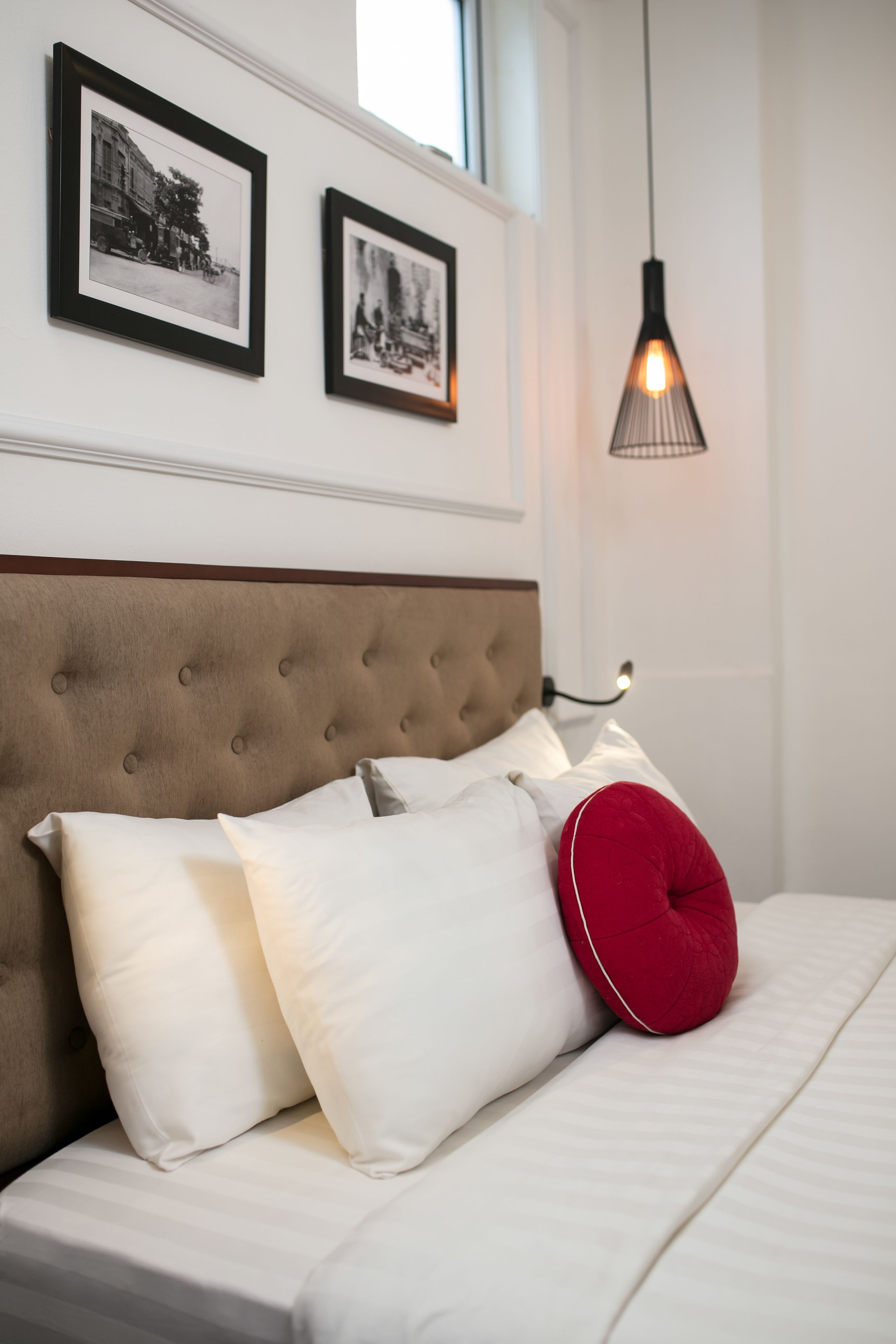 A neatly made bed with white and red pillows, a tufted headboard, and two framed photographs on the wall above.