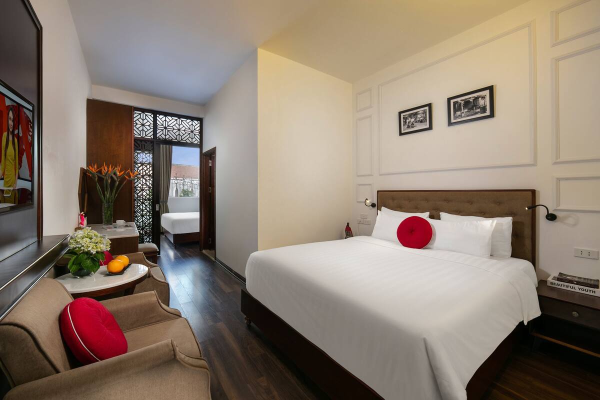 A hotel room with a large bed, a small sitting area with a red cushion, a vase with flowers, and a view of the outside.
