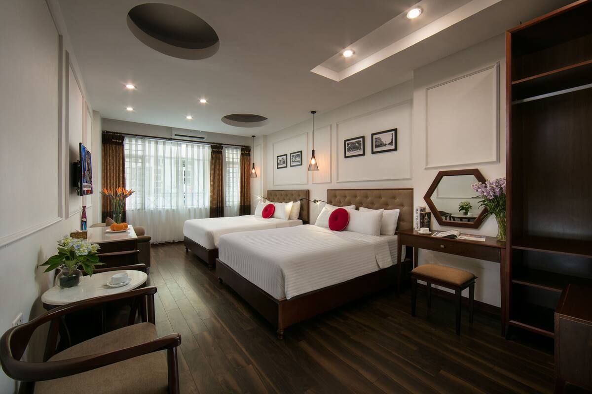 A hotel room with two beds, a desk, a chair, a small table with a vase of flowers, and a mirror.