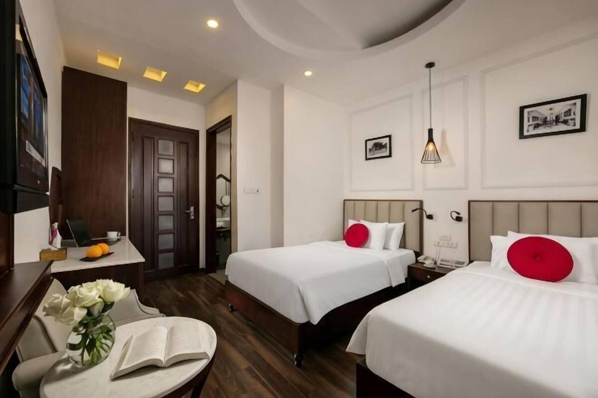 A hotel room with two beds, a desk with a chair, a small table with a vase and book, and a door leading to another room.