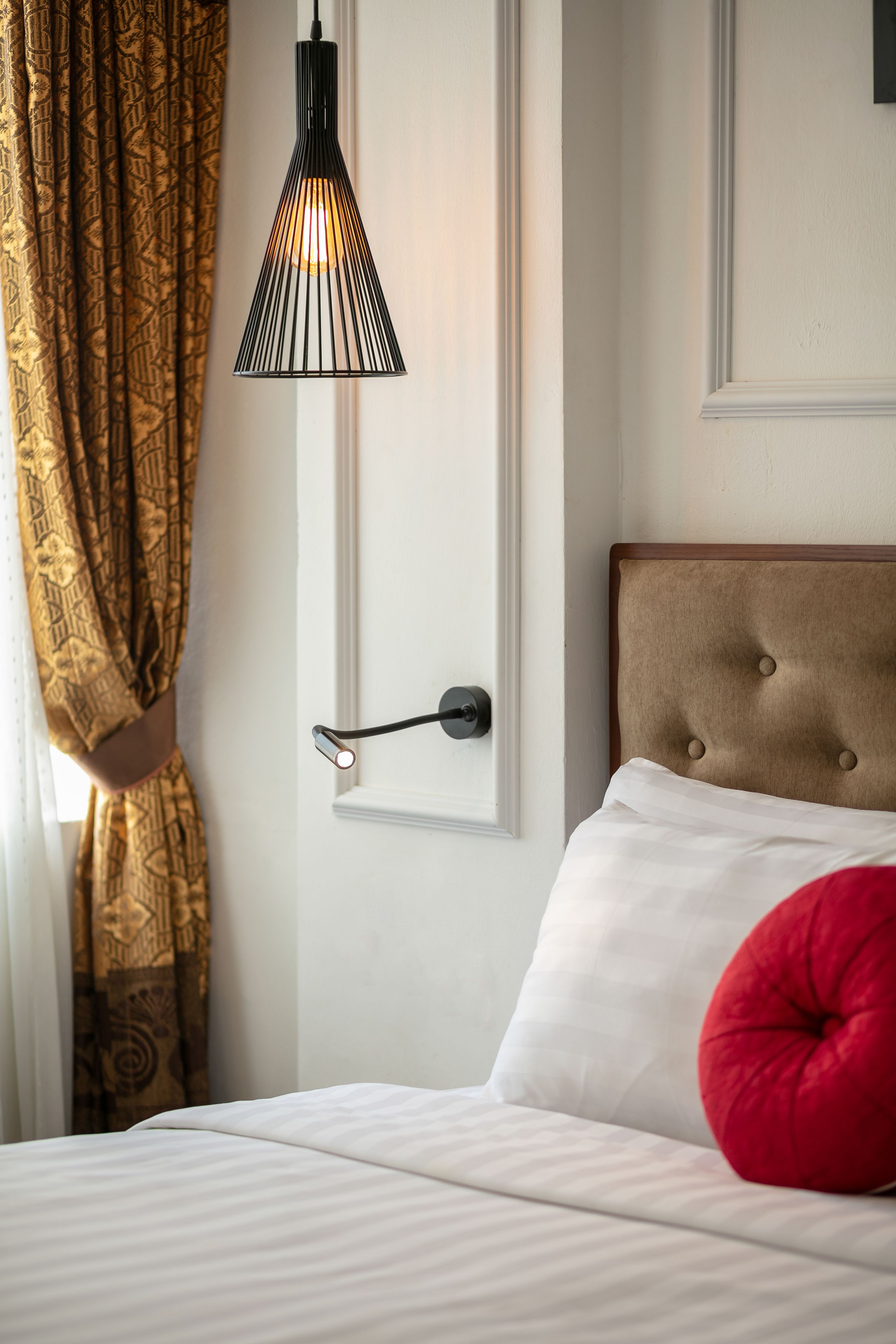 A hotel room with a bed, a red pillow, a black pendant light, and patterned curtains.