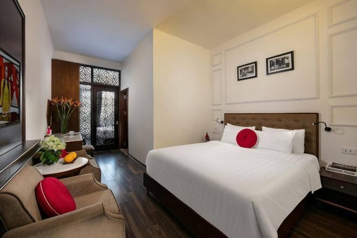 A hotel room with a large bed, a small table with a vase and fruit, a chair, and a decorative partition.