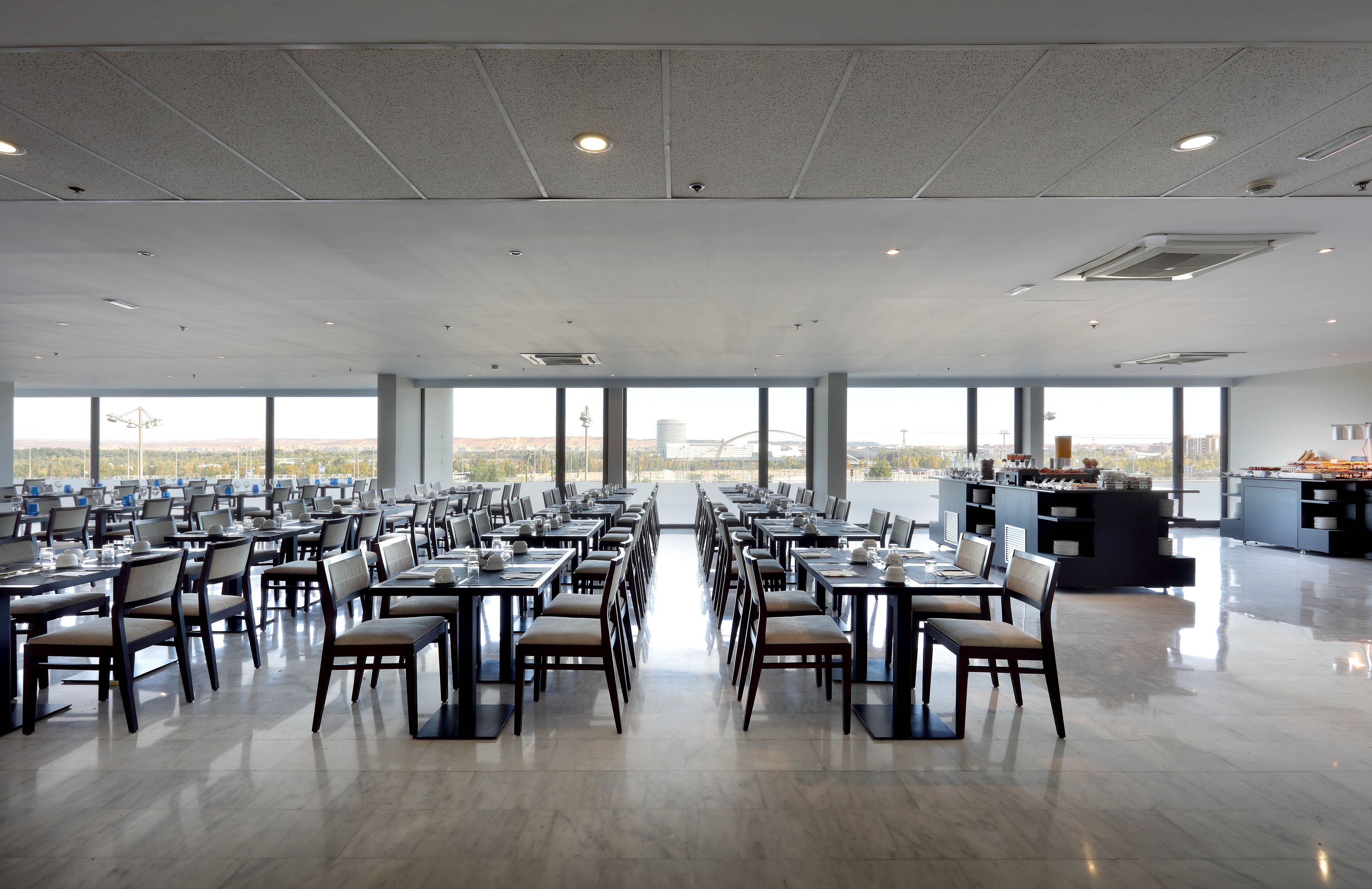 A spacious dining area with multiple tables and chairs, large windows offering a view of the cityscape, and a kitchen area visible in the background.