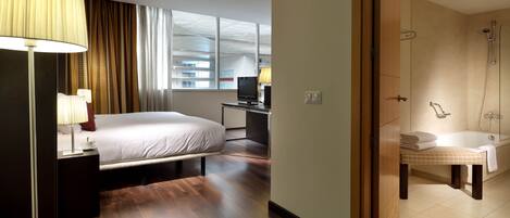 A hotel room with a bed, bedside table, lamp, desk, computer, and a bathroom with a sink and shower.