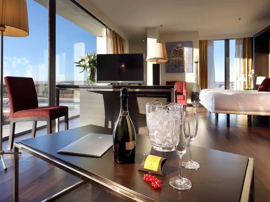 Habitación de hotel con una televisión grande, una cama, un escritorio con una computadora portátil, una botella de vino y un jarro de hielo.