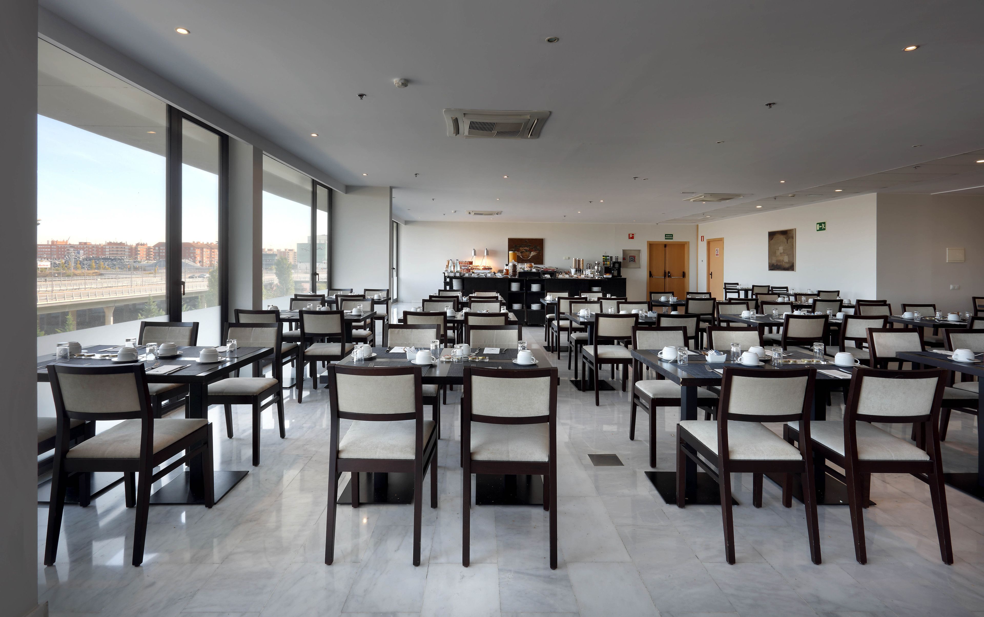 Un comedor amplio con varias mesas y sillas, ventanas grandes que ofrecen vista a la ciudad, y un diseño moderno en el techo.