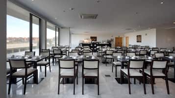 Un comedor amplio con varias mesas y sillas, ventanas grandes que ofrecen vista a la ciudad, y un diseño moderno en el techo.