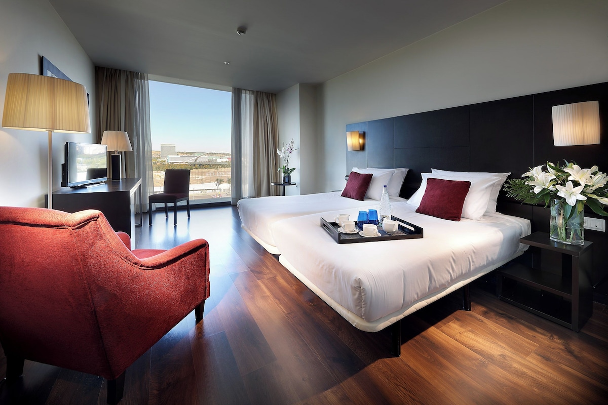 Een moderne hotelkamer met een groot bed, een bureau, een rode fauteuil en uitzicht op de stad.