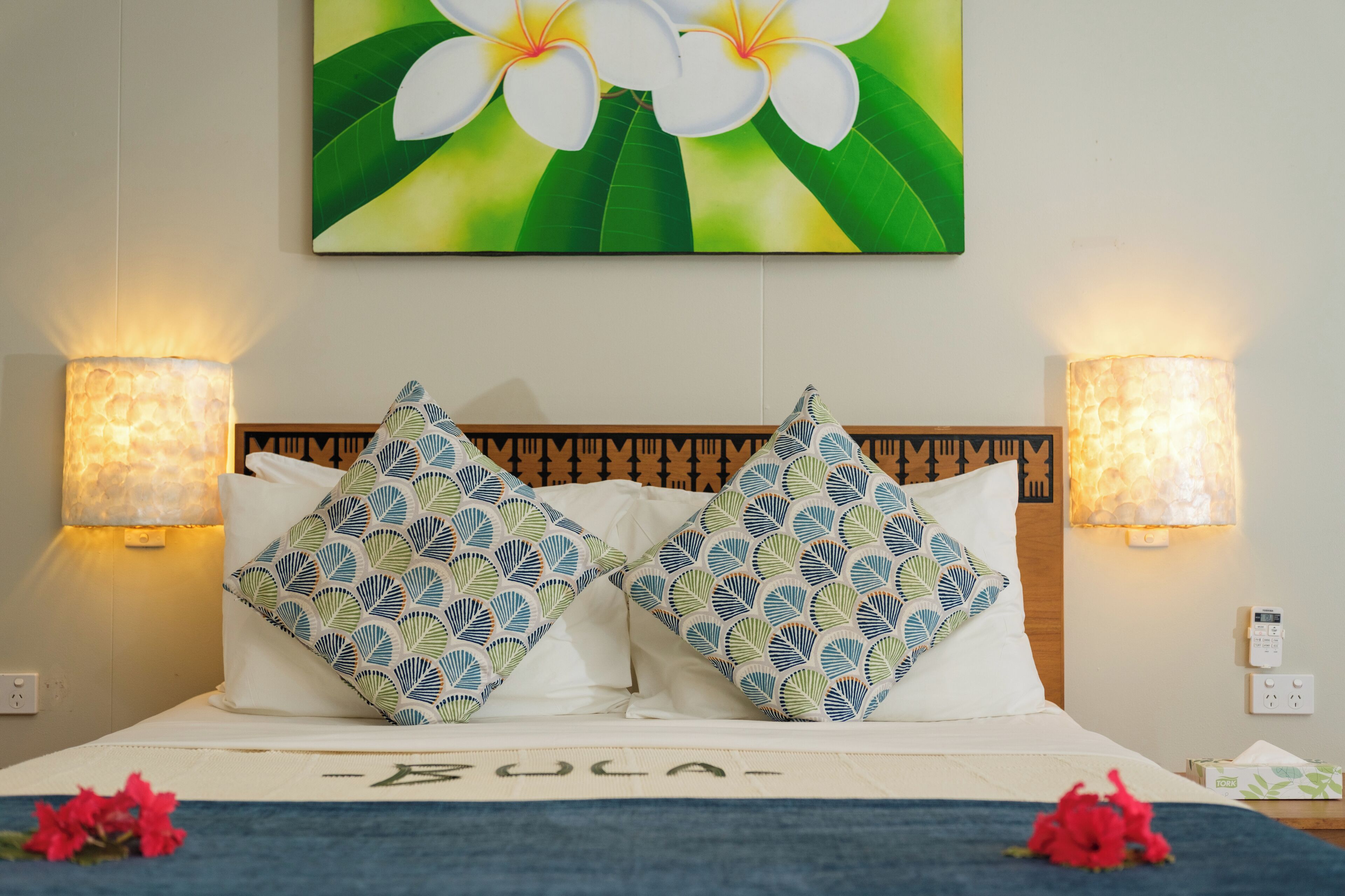 A hotel room with a bed, two patterned pillows, a wall-mounted painting of flowers, and two wall-mounted lamps.