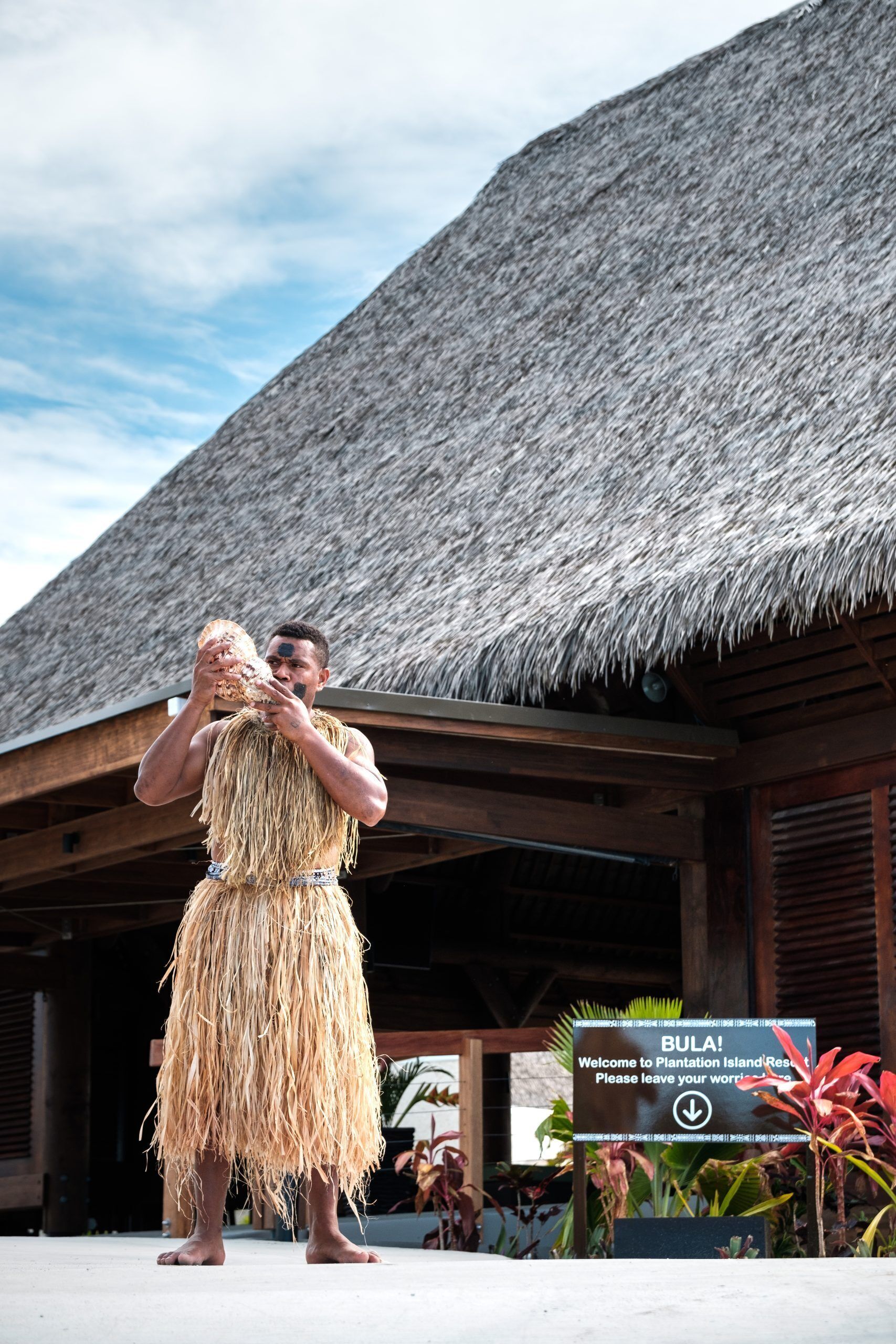 A person in a straw skirt standing in front of a thatched-roof building with a sign that reads "Welcome to Plantation Island Resort."