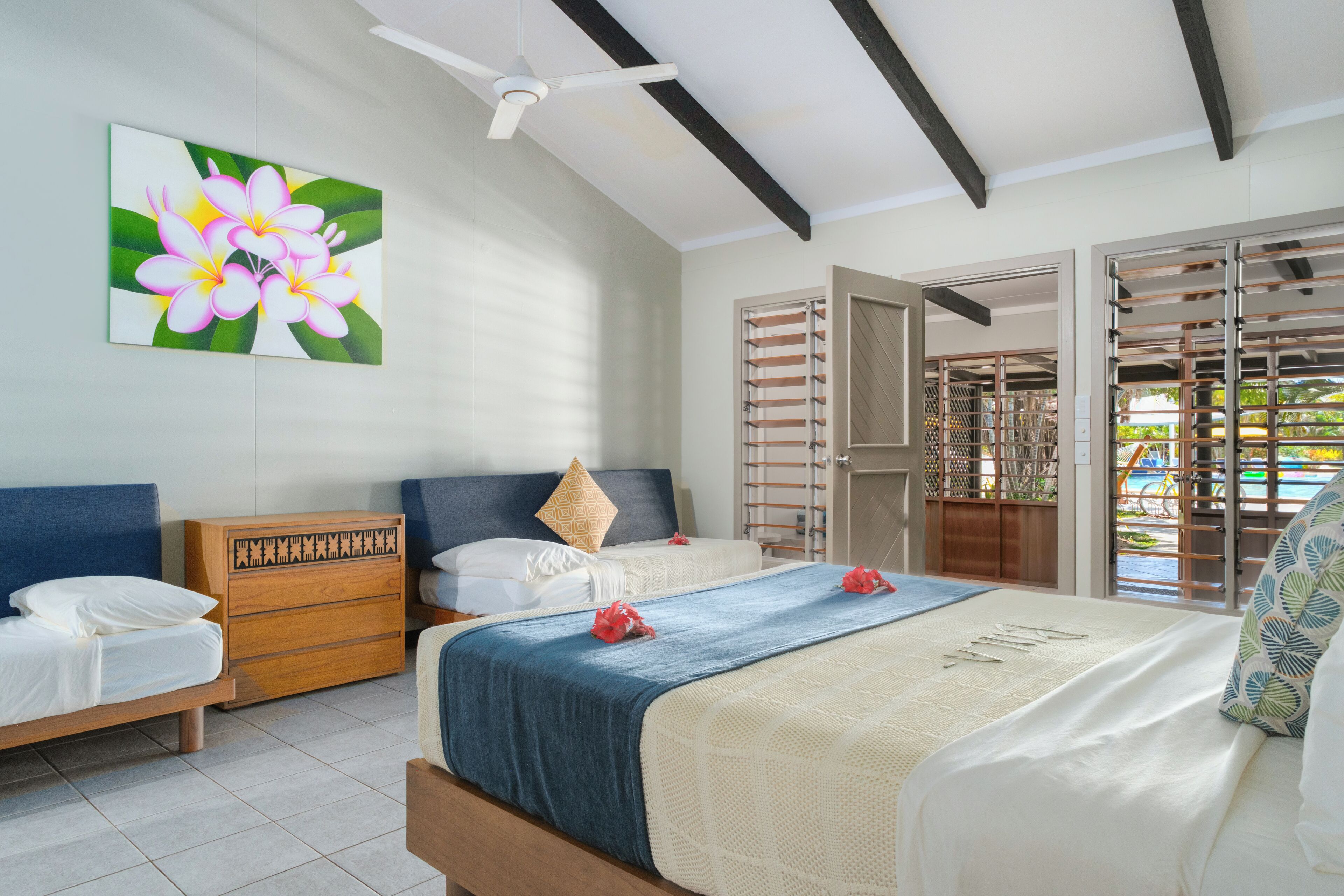 Une chambre avec deux lits, une commode en bois, un ventilateur de plafond et une grande fenêtre donnant sur une piscine.