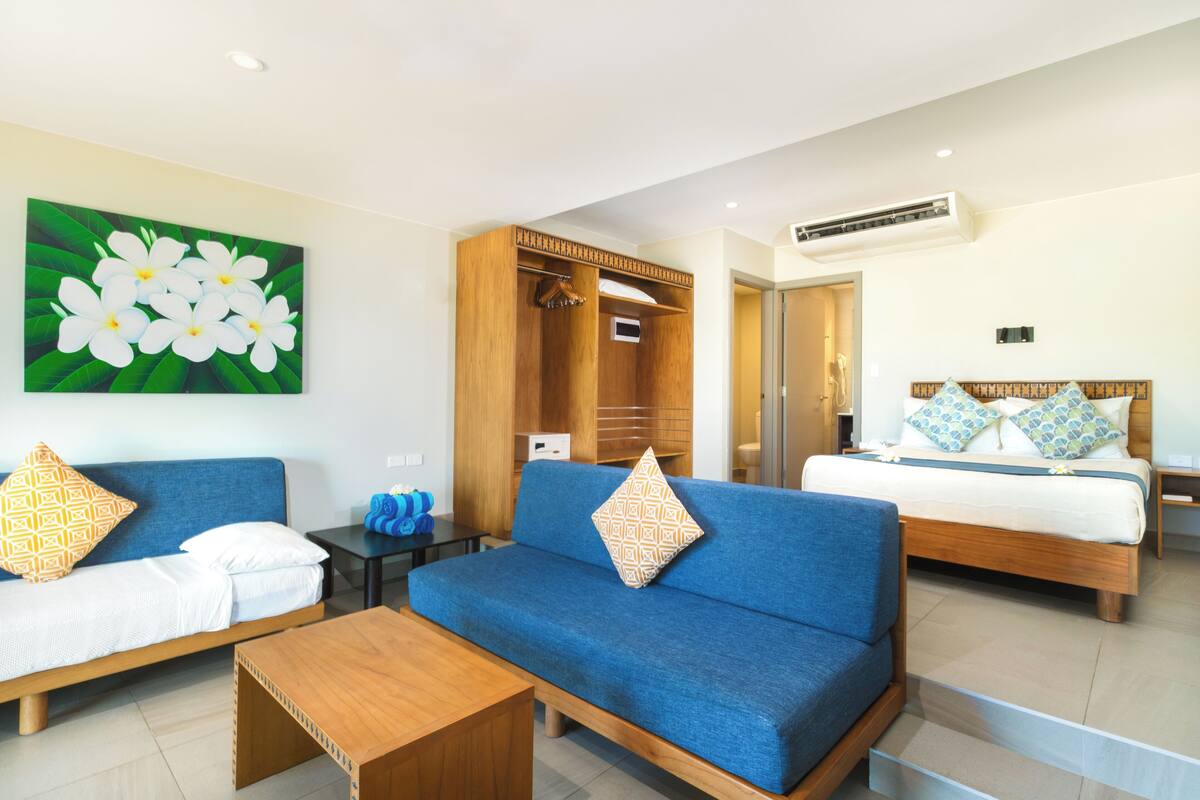 A hotel room with a blue sofa, a wooden coffee table, a bed with a wooden headboard, and a wardrobe.