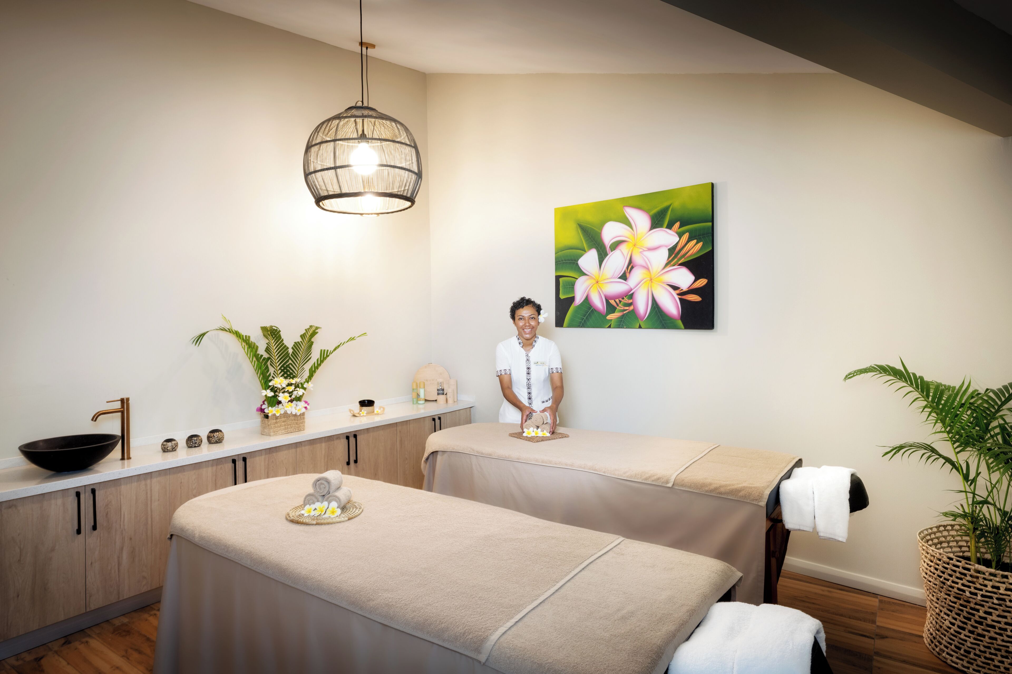 A spa room with two massage tables, a sink, a large window, and a painting of flowers on the wall.