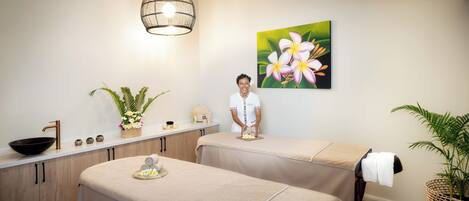 A spa room with two massage tables, a sink, a large window, and a painting of flowers on the wall.