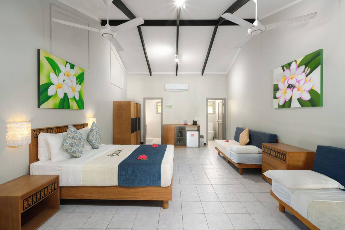 A hotel room with a large bed, two smaller beds, a wooden dresser, and a desk. There are two paintings on the walls, a ceiling fan, and an air conditioning unit.