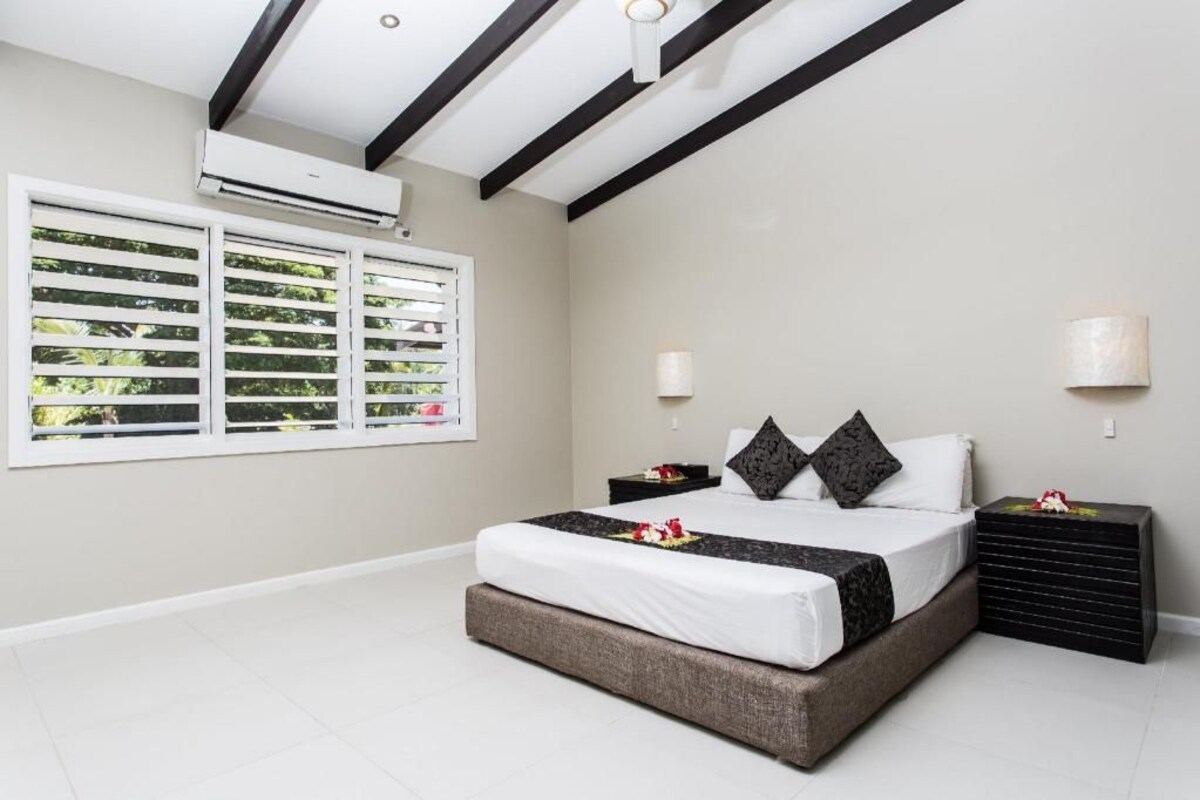 Un dormitorio con cama, dos mesitas de noche, ventana con persianas, unidad de aire acondicionado y techo con vigas negras.