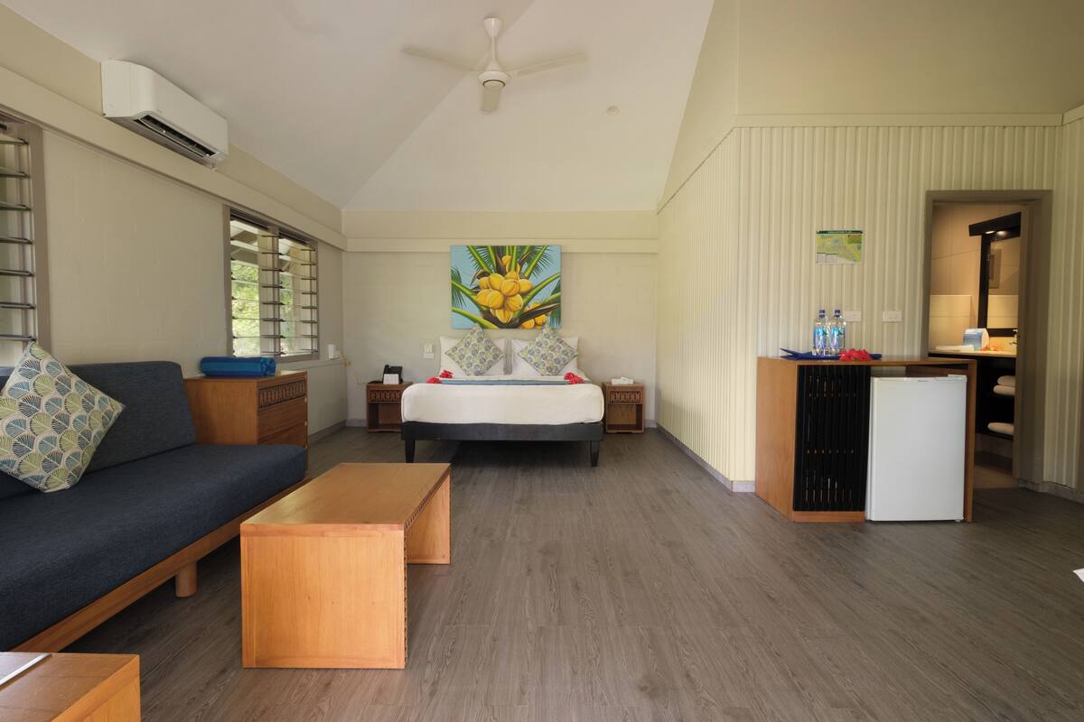 A spacious room with a bed, a sofa, a small wooden table, a ceiling fan, and a refrigerator.