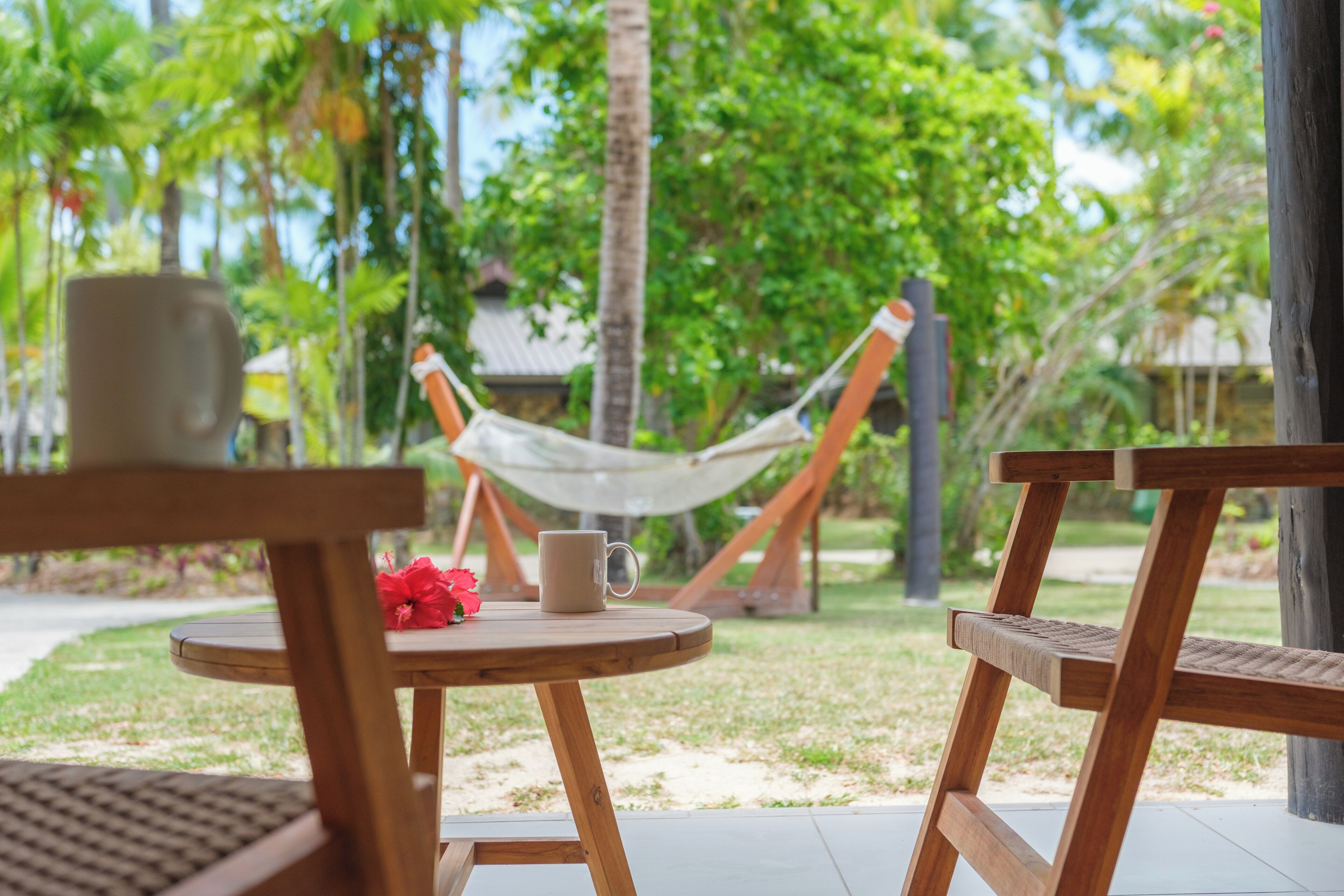 A wooden table with a cup and a red flower, a hammock, and a view of a garden with trees and a building in the background.