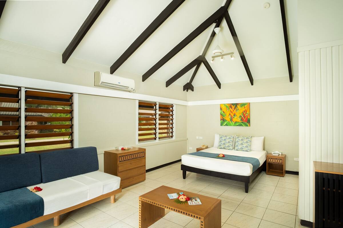 A room with a bed, a sofa, a coffee table, a dresser, and a window with blinds.