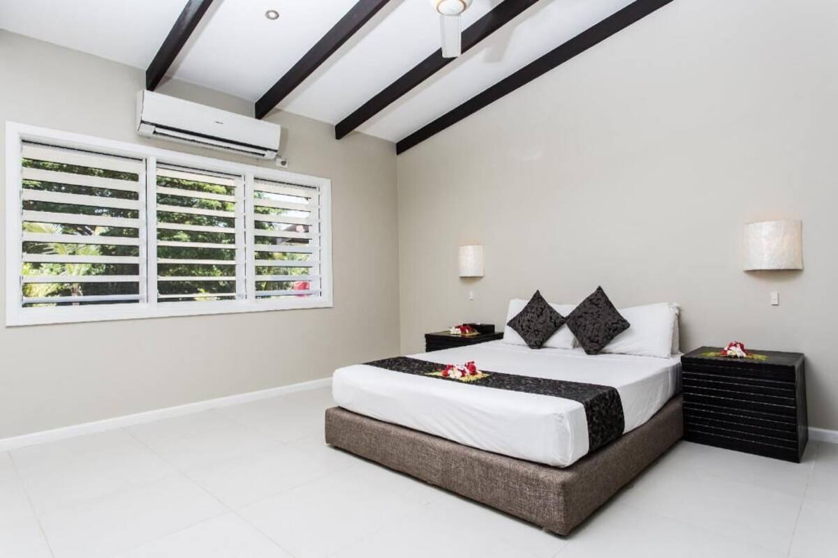 A bedroom with a bed, two bedside tables, a window with blinds, an air conditioning unit, and a ceiling with black beams.