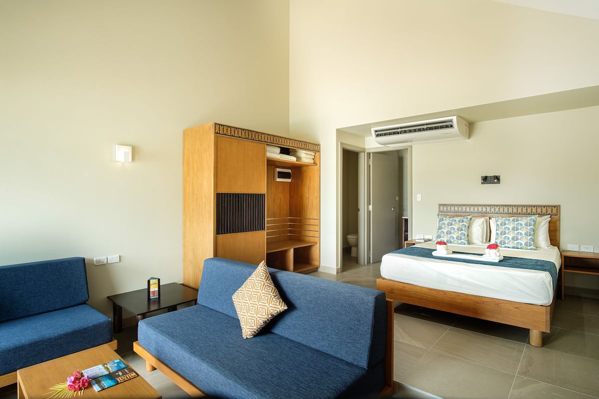 A hotel room with a bed, a sofa, a small table, a wardrobe, and an air conditioning unit.