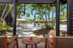 Un espacio de estar al aire libre techado con vista a la piscina, muebles de madera y plantas tropicales.