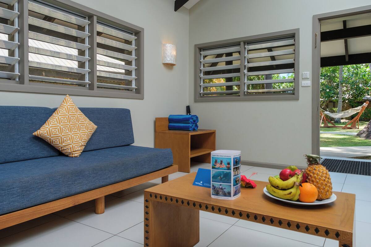 A room with a blue sofa, a wooden coffee table with a fruit plate, and a window with blinds.