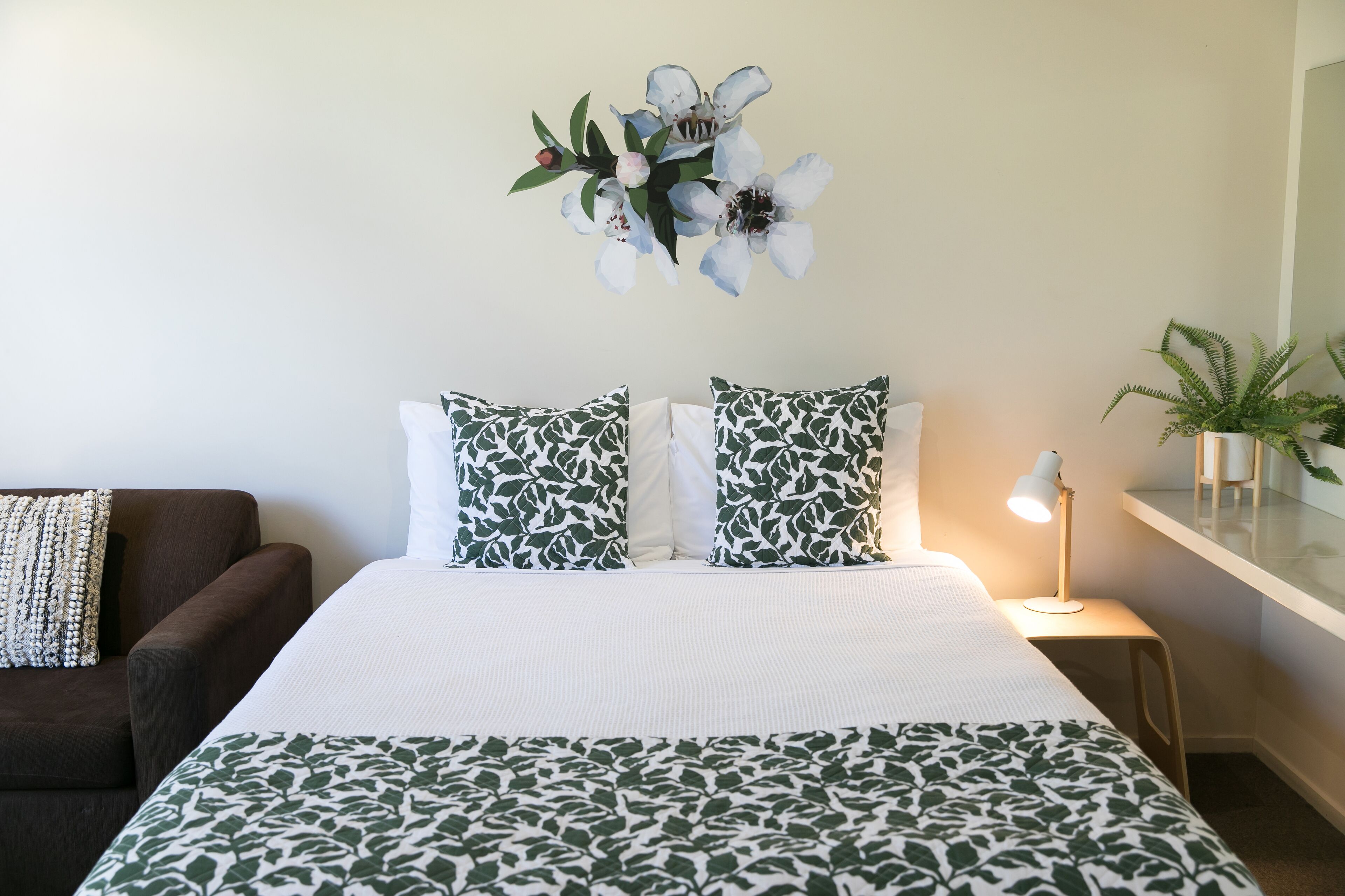 Een netjes ingerichte slaapkamer met een bed, kussens, een nachtkastje, een lamp en een wandplant.