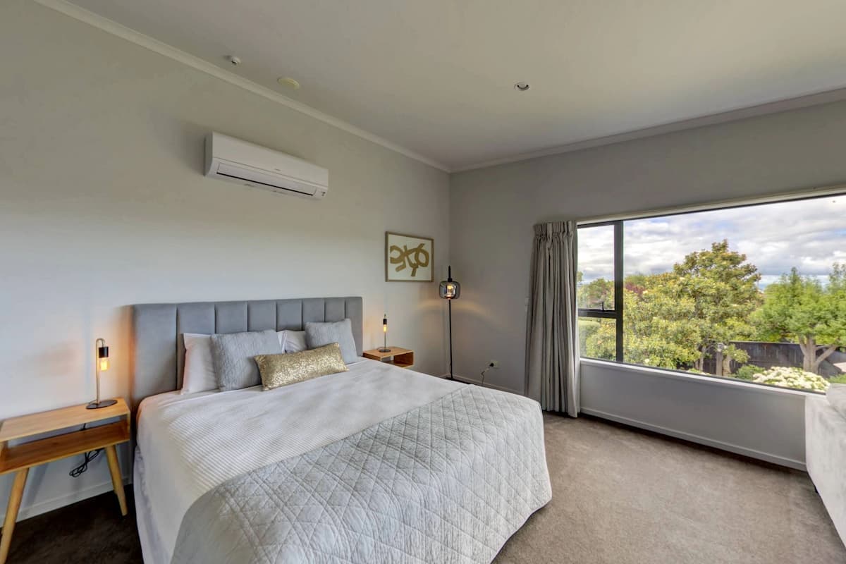 Un dormitorio moderno con una cama grande, una mesita de noche con lámpara, una ventana con vistas a árboles y un aire acondicionado instalado en el techo.