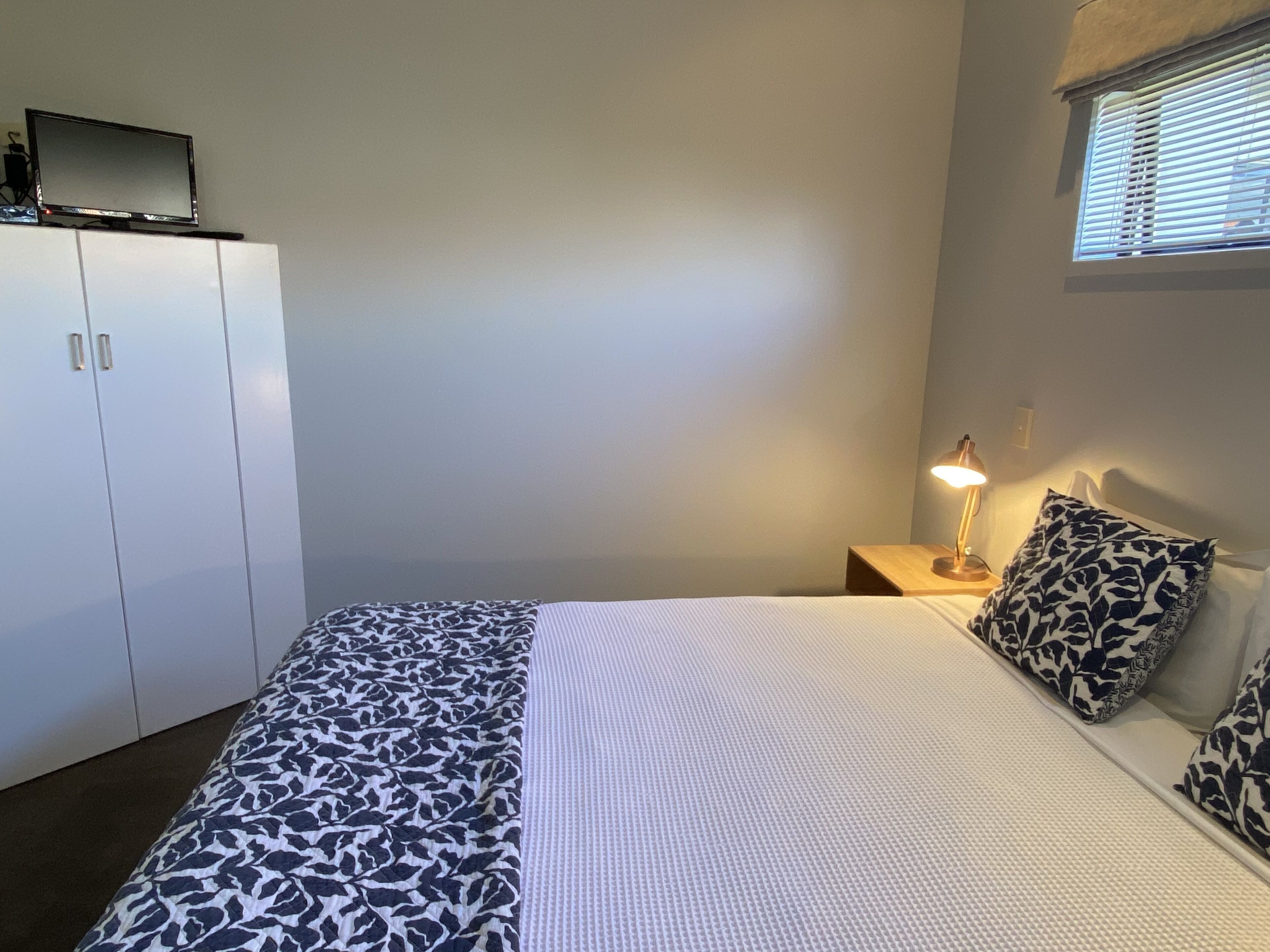 Un dormitorio con cama, mesita de noche con lámpara, armario y ventana con persianas.