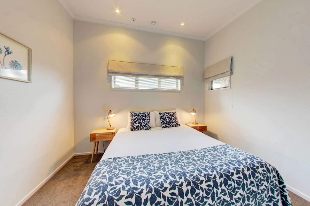 Un dormitorio con cama, lámparas de noche, una ventana con persianas, un cuadro en la pared y una lámpara de techo.