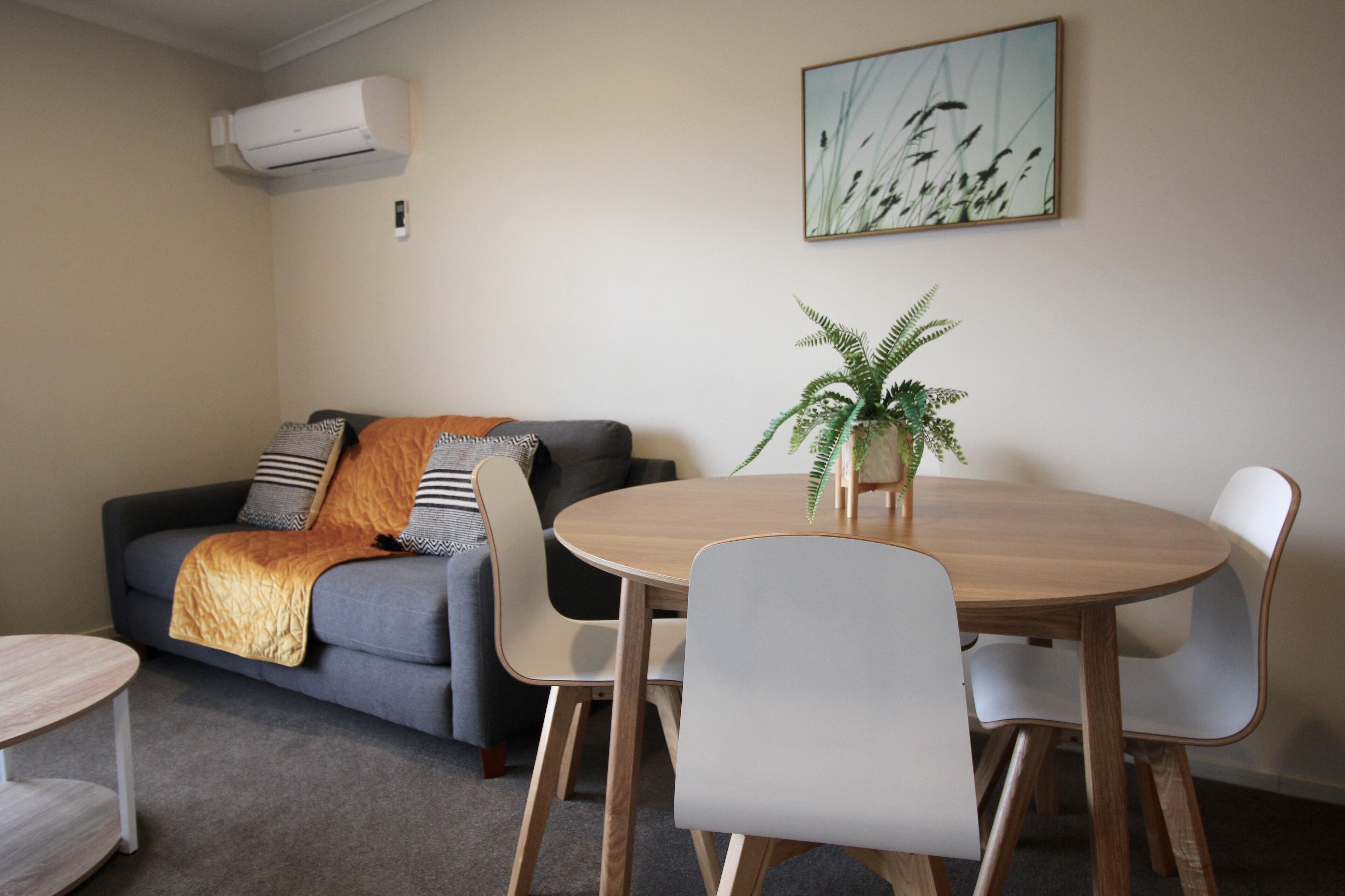 Un salon avec un canapé gris, une table ronde en bois et des chaises blanches. On peut voir une plante en pot sur la table et un tableau au mur.